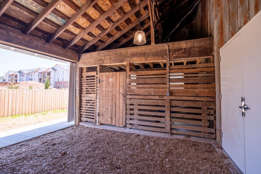 A covered stable breezeway with slatted wooden partitions, a pendant light overhead and an open bay looking out to the fenced yard.