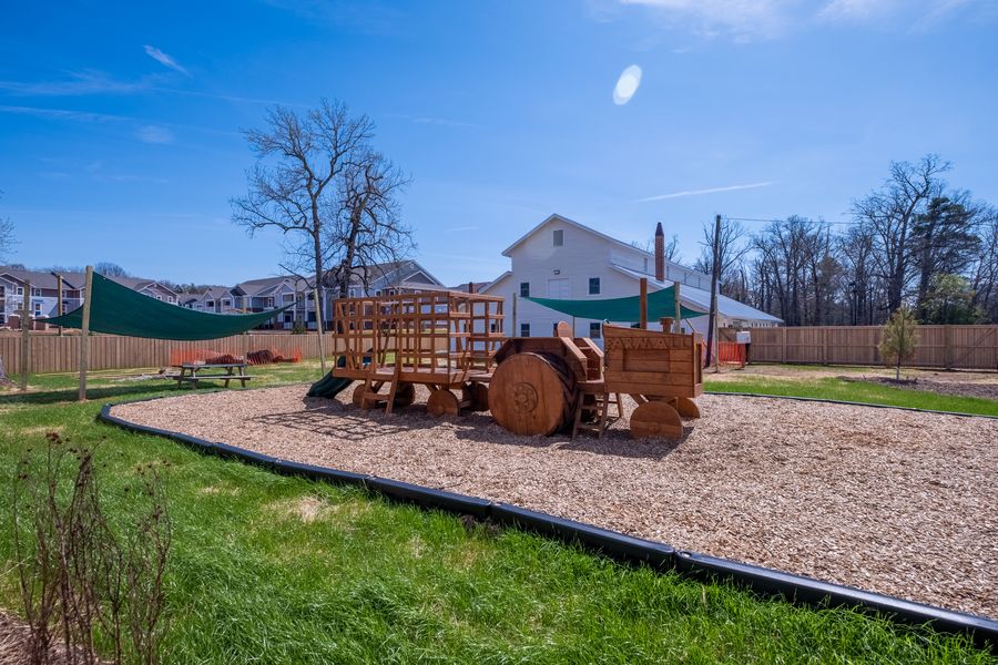 A wider view of the play yard: mulch surfacing edged in black, picnic tables on the grass, shade sails on timber posts and the barn beyond.