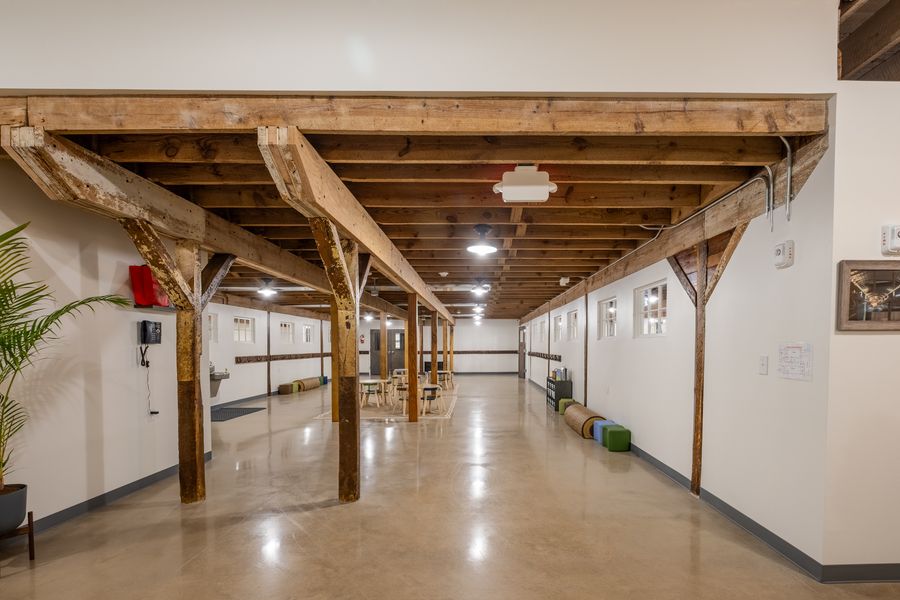 A long open common room under exposed timber beams and posts, with coat hooks along the wall, a drinking fountain and low tables at the far end.
