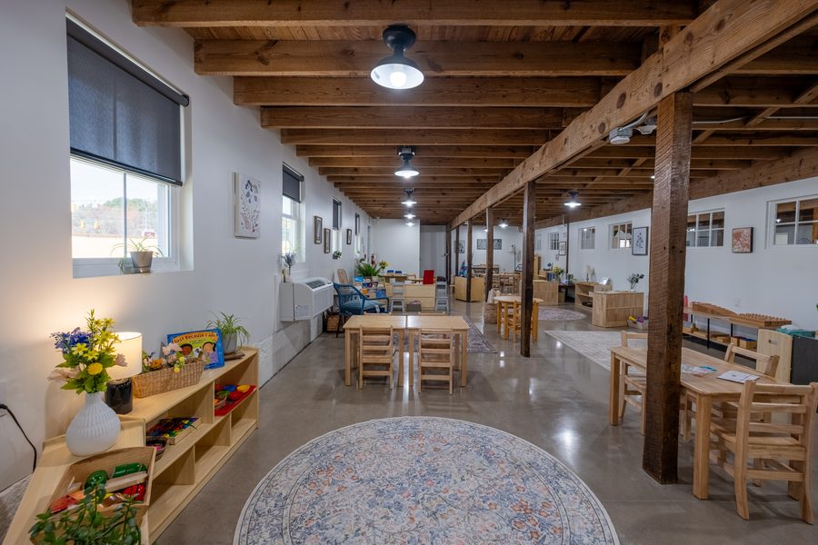 A long view of the Primary classroom with a round patterned rug in the foreground, tables set for work and shelving along both walls.