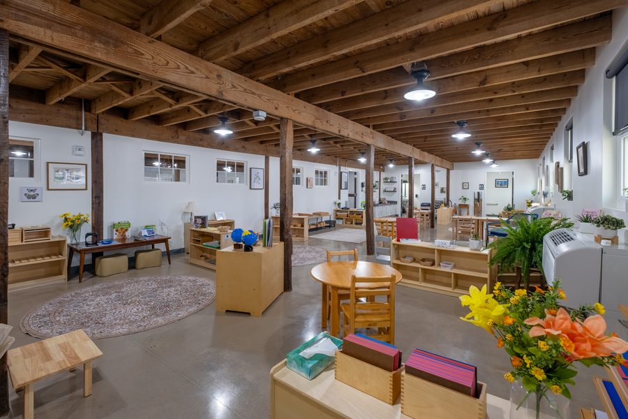The Primary classroom with a round rug, low shelving of materials, globes on a stand and flowers on the counter beside the window.