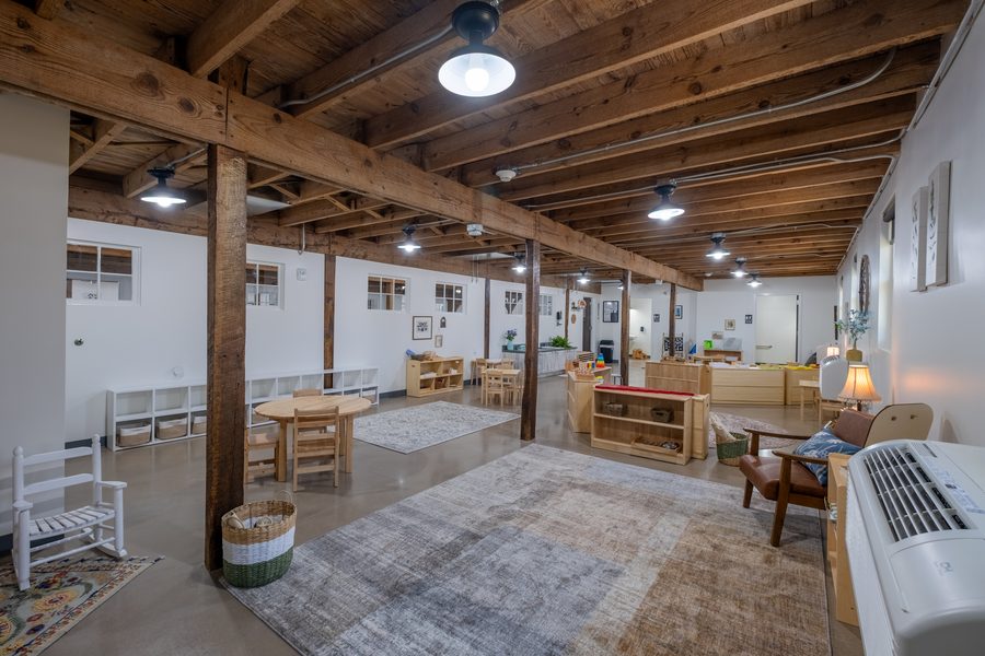 A wide view of the Toddler classroom with a white rocking chair, low cube storage with baskets, a round table and layered rugs on polished concrete.